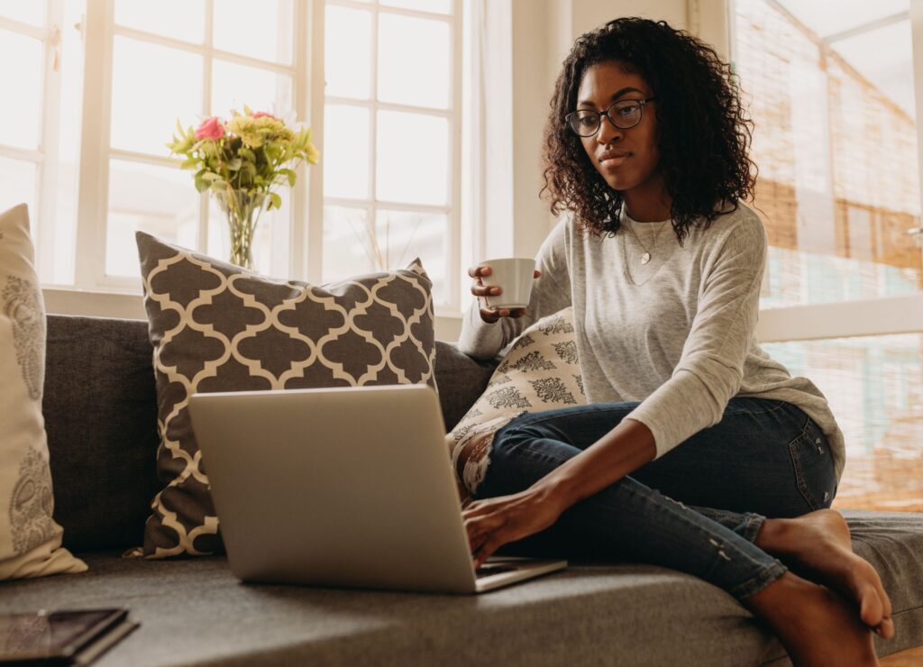 Businesswoman working on laptop computer sitting at home holding a coffee cup in hand. Woman sitting on sofa at home and managing her business.